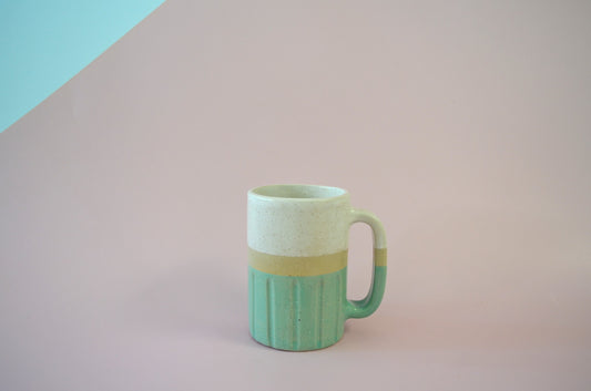 Picture of a handmade mug with the top half and interior painted white and the bottom half painted light green. There is a carved patten of thick vertical lines along the bottom half.
