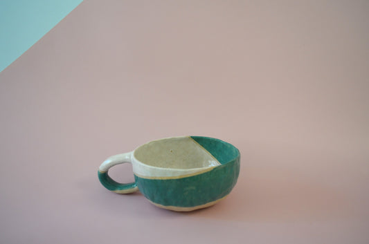 Picture of a handmade bowl shaped mug with a protruding crescent shaped handle, that is half painted in white and the other half painted in teal.